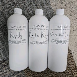 Puede incluir: Tres botellas de plástico blanco de detergente líquido para la ropa con etiquetas negras. Las etiquetas dicen "R&W CO. 32oz Liquid Laundry Detergent" y tienen los nombres "Royalty", "Belle Rouge" y "Sun Washed Linen" impresos en ellas.
