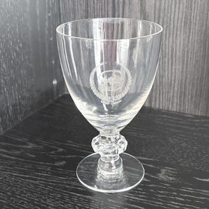 Puede incluir: Una copa transparente con un emblema decorativo grabado en el cuenco. La copa tiene una base redonda y un tallo facetado. Adecuada para servir bebidas.