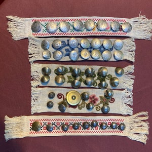 May include: Five rows of decorative metal buttons with different designs and finishes, sewn onto strips of white fabric with embroidered floral patterns. The buttons are silver, gold, and brass.