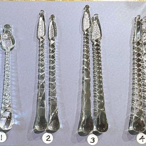 May include: Four pairs of clear glass serving spoons with decorative stems and spoon heads. The spoons have different designs, including twisted and textured patterns. The spoons are arranged on a light gray surface.