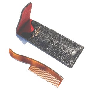 May include: A tortoiseshell comb with a curved handle rests beside a black leather case with a red interior. The case is embossed with gold lettering. The comb has fine teeth and a smooth, polished finish.