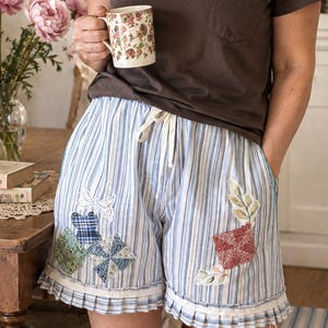 May include: Light blue and white striped shorts featuring a drawstring waist, a ruffled hem, and various fabric patches including a white lace butterfly, green and blue checkered squares, and a red patterned square with leaf accents.