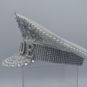 Pode incluir: Um chap&eacute;u de capit&atilde;o branco com enfeites de p&eacute;rolas e strass. O chap&eacute;u tem uma faixa prateada com strass e a palavra "LOVE" escrita em strass na frente.