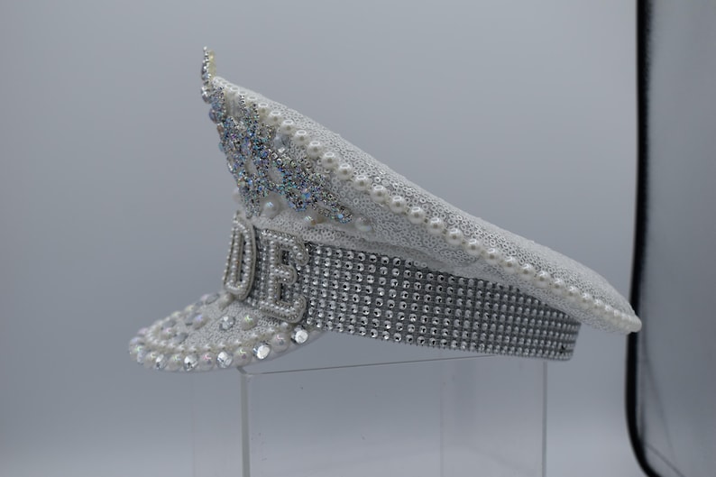 Pode incluir: Um chap&eacute;u de capit&atilde;o branco com enfeites de p&eacute;rolas e strass. O chap&eacute;u tem uma faixa prateada com strass e a palavra "LOVE" escrita em strass na frente.