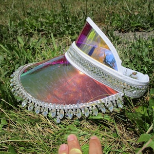 May include: A clear iridescent visor with a white trim and a row of rhinestones around the brim. The visor is lying on a bed of green grass.