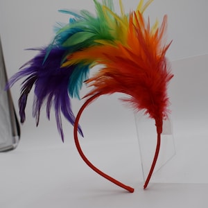 May include: A rainbow feather headband with a red headband. The feathers are arranged in a rainbow pattern, starting with purple at the bottom and ending with yellow at the top.