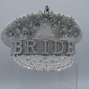 Pode incluir: Chap&eacute;u de noiva branco com uma coroa de strass e p&eacute;rolas. A palavra "BRIDE" &eacute; escrita em strass na frente do chap&eacute;u.