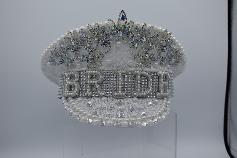 Pode incluir: Chap&eacute;u de noiva branco com uma coroa de strass e p&eacute;rolas. A palavra "BRIDE" &eacute; escrita em strass na frente do chap&eacute;u.
