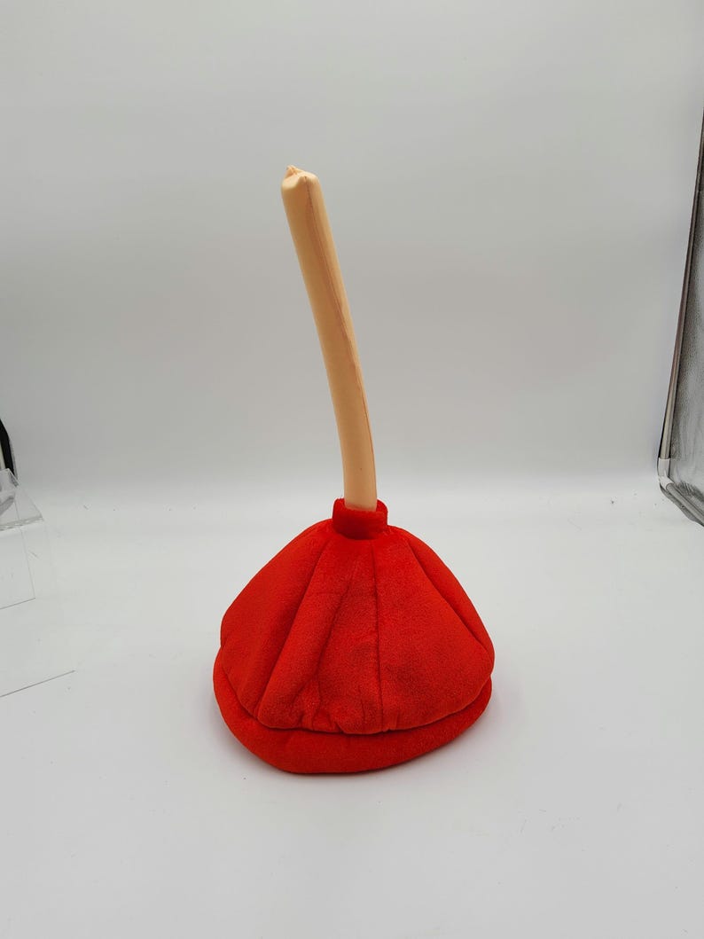 May include: A red plush plunger with a brown handle. The plunger is shaped like a traditional toilet plunger with a round, flat bottom and a long, thin handle.