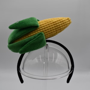 May include: A novelty headband featuring a plush corn on the cob design. The corn is yellow with green husks, attached to a black headband. This playful accessory is suitable for costume parties or themed events.