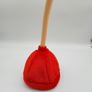 May include: A red plush plunger hat with a long, cream-colored handle. The hat is shaped like a traditional plunger with a rounded top and a flat base.