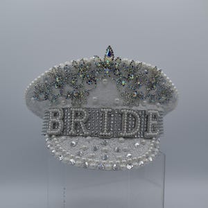 Pode incluir: Chap&eacute;u de noiva branco decorado com p&eacute;rolas e strass. A palavra "BRIDE" &eacute; escrita em strass na frente do chap&eacute;u.