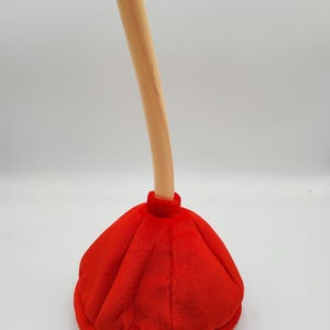 May include: A red and beige plush plunger hat. The hat is shaped like a plunger with a long, thin handle.