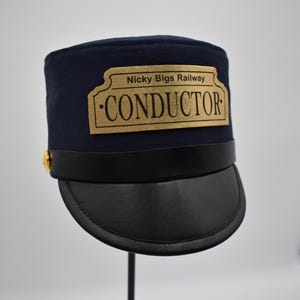 May include: A blue conductor's hat with a black brim and a gold badge that reads "Nicky Bigs Railway - CONDUCTOR".