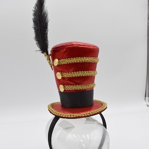 May include: A red and gold top hat headband with a black feather. The hat has three gold bands and gold buttons. The headband is black.