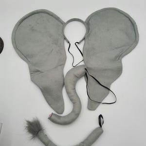 May include: Gray elephant costume set with a headband, ears, and tail. The ears are made of soft, plush fabric and are attached to a headband. The tail is also made of plush fabric and has a fluffy tip.