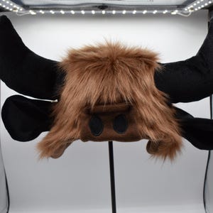 May include: A novelty hat shaped like a bull's head. The hat features large black horns and ears, with a brown faux fur mane and a brown face with two black eye cutouts. The hat is designed to be worn on the head.