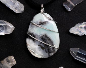 Peut inclure: Un pendentif en forme de goutte composé d'une pierre blanche et vert menthe avec des inclusions gris foncé, entouré de fil métallique argenté et suspendu à un cordon noir, entouré de huit pointes de cristal de quartz clair.