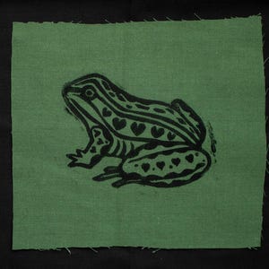 May include: A square, green fabric patch featuring a black frog illustration. The frog has heart-shaped markings and striped details. The fabric has a raw, unfinished edge, and the background is black.