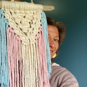 May include: A macrame wall hanging with a wooden dowel at the top. The hanging features a geometric pattern with a white, pink, and light blue color scheme.