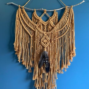 May include: A macrame wall hanging with a natural wood branch at the top. The macrame is made with a thick, natural-colored cord and features a geometric pattern. Two black feathers are hanging from the center of the macrame.