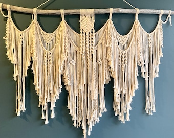May include: A large macrame wall hanging with a natural beige color. The hanging is made of cotton cord and has a geometric pattern. It is hung on a wooden branch.