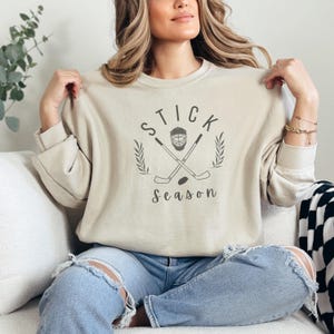 May include: A beige sweatshirt with the text "STICK SEASON" in a black font. A hockey stick and a hockey mask are crossed in the center of the text.