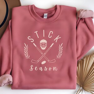 May include: A pink sweatshirt with the text "STICK SEASON" in white. The design includes a hockey stick, a hockey puck, and a hockey helmet.