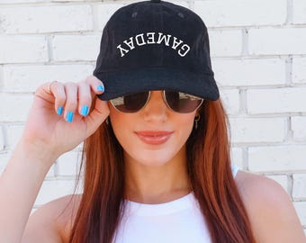 Könnte beinhalten: Schwarze Baseballkappe mit dem Wort "GAMEDAY" in Weiß, das sich über die Vorderseite wölbt. Die Kappe wird von einer Person getragen, die eine Sonnenbrille und ein weißes Oberteil trägt. Der Hintergrund ist eine weiße Backsteinmauer.