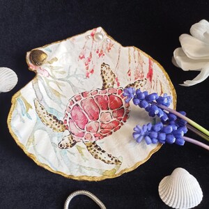 Könnte beinhalten: Dekorative Muschelschalen-Schmuckschale mit handgemaltem Schildkröten-Design. Die Schale hat einen goldfarbenen Rand und ist mit Perlen und blauen Blumen verziert. Der Schildkrötenpanzer ist rot und rosa. Der Hintergrund ist schwarz.