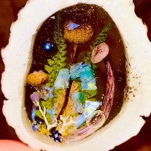 Op de afbeelding: Handgemaakte hanger met een miniatuurlandschap ingekapseld in heldere hars. De hanger toont gedroogde bloemen, paddenstoelen en iriserende accenten in een wit, organisch gevormd frame. Het is opgehangen aan een zilverkleurige beugel.