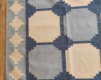 May include: A woven rug with a geometric pattern in shades of blue and cream. The rug features a repeating design of squares and crosses, creating a textured surface. The border is a lighter blue with cream-colored crosses.