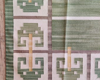 May include: A green and white patterned rug with a geometric design. The rug features repeating square patterns with green and gold accents, and a fringed edge. The rug has a striped section on the right side.