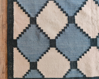 May include: A woven rug with a geometric pattern. The rug features a repeating design of blue, off-white, and dark blue diamond shapes. The border is a solid dark blue. The rug is made of a textured material.
