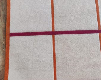 Op de afbeelding: Een wit tapijt met een rasterpatroon in oranje en bordeauxrood. Het tapijt heeft een dikke, pluche textuur.