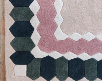 May include: A close-up of a geometric rug with a soft, textured surface. The rug features a light pink background with a pink, white, and dark green hexagonal pattern border. The rug's design is modern and stylish.