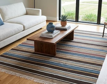 Op de afbeelding: Een rechthoekig vloerkleed met een horizontaal streeppatroon in tinten bruin, blauw, beige en wit. Het kleed heeft een platgeweven textuur en is voorzien van korte, lichtgekleurde franjes langs de twee kortere zijden van het textiel.