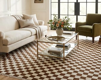 May include: A rectangular area rug featuring a repeating checkerboard pattern in alternating dark brown and cream squares. The rug has a textured, woven pile and short fringe detailing along the edges, covering a light-coloured wooden floor.