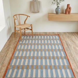 Op de afbeelding: Een rechthoekig vloerkleed met een herhalend geometrisch patroon van verticale lichtblauwe en beige strepen op een crèmekleurige achtergrond, afgewerkt met een effen roestoranje rand die de gehele omtrek van het kleed omlijst.