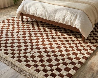 May include: A brown and cream checkered rug with a fringed edge is placed on a wooden floor. The rug is under a bed with a wooden frame, white bedding, and a beige blanket. A nightstand with a lamp and a plant are visible.