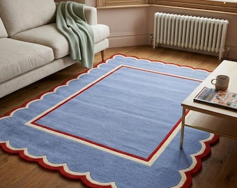 May include: A rectangular area rug featuring a solid light blue center, a cream-colored border, and a scalloped edge finished in a bold red trim. The rug is laid flat on a wooden floor, showcasing a clean design with distinct color-blocked sections.