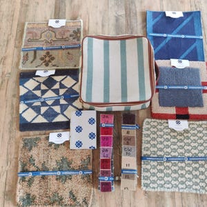 Puede incluir: Una variedad de muestras de alfombras y una bolsa a rayas se exhiben sobre una superficie de madera. Las muestras presentan diversos patrones y colores, incluyendo diseños geométricos, motivos florales y texturas lisas. También se incluye una tarjeta de muestras de color.