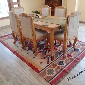 May include: A wooden dining table with six chairs, set with a green table runner and placemats. The table and chairs are in a light wood tone. The table is set on a rug with a geometric pattern in shades of red, brown, and beige.