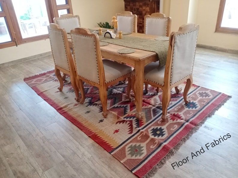 May include: A wooden dining table with six chairs, set with a green table runner and placemats. The table and chairs are in a light wood tone. The table is set on a rug with a geometric pattern in shades of red, brown, and beige.