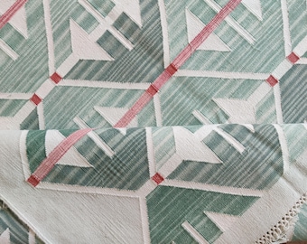 May include: A folded green and white patterned textile with a geometric design. The pattern includes white arrow-like shapes, diagonal lines, and red accents. The edge of the textile has a fringe.