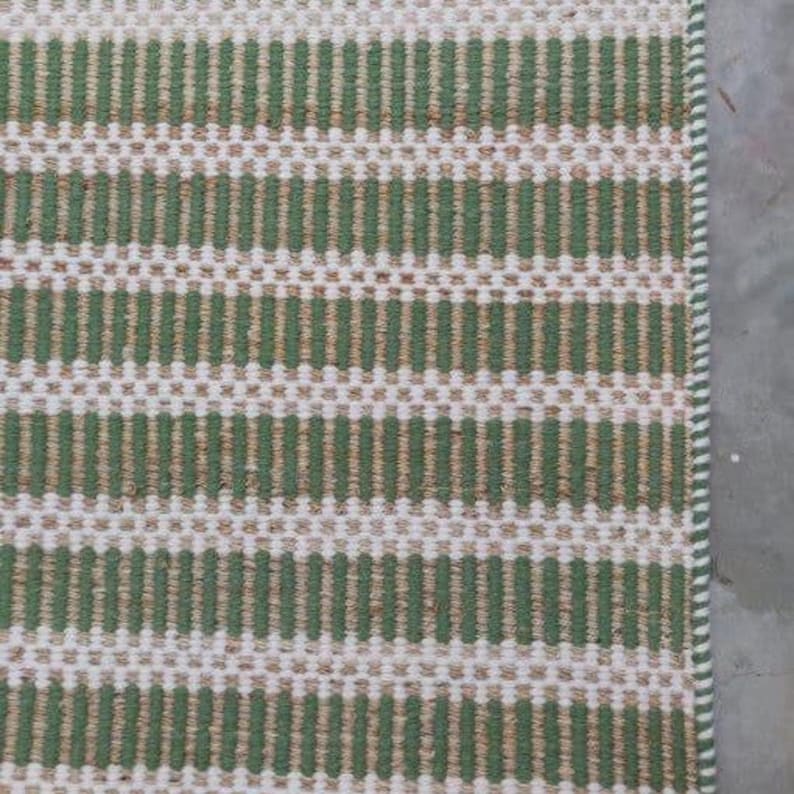 May include: A green and white striped woven rug with a subtle texture. The rug has a simple design and is made of natural fibers.