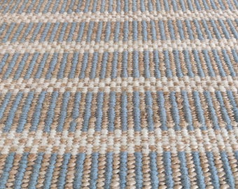 Puede incluir: Primer plano de una alfombra tejida con un patrón de rayas. La alfombra presenta bandas alternas de azul claro, beige y blanco. Las patas de una silla de madera son visibles en la parte superior de la imagen.