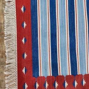 May include: A red, blue, and white striped rug with a fringe border. The rug has a geometric pattern in the border.