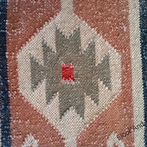 May include: Close-up of a woven rug with a geometric pattern in shades of red, brown, white, and gray. The pattern features a central diamond shape with a smaller red diamond in the center.