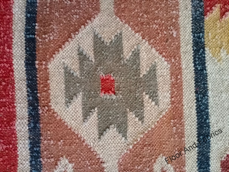 May include: Close-up of a woven rug with a geometric pattern in shades of red, brown, white, and gray. The pattern features a central diamond shape with a smaller red diamond in the center.